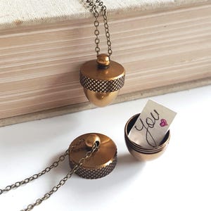 May include: A bronze-colored acorn pendant necklace with a chain. The acorn pendant is open, revealing a small note that says "You" with a heart. The acorn has a textured design.