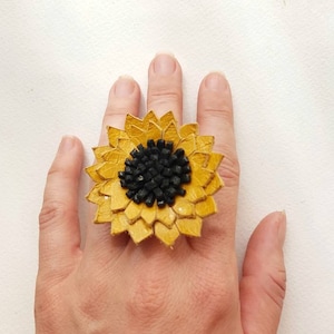 Puede incluir: Un anillo de girasol amarillo con centro negro, usado en una mano. Los pétalos del anillo están superpuestos, creando un efecto tridimensional. El anillo es una pieza de joyería única.