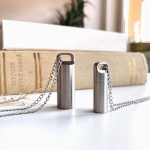 May include: Two cylindrical silver-toned metal cremation urn pendants with a brushed finish. Each pendant features a small loop at the top attached to a delicate silver-toned metal chain. The pendants are standing upright on a flat surface.