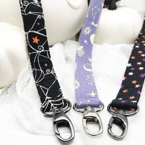 May include: Three fabric lanyards with metal swivel clasps. The lanyards feature different patterns: a black one with white spiderwebs and orange stars, a purple one with celestial designs, and a black one with orange and purple stars.