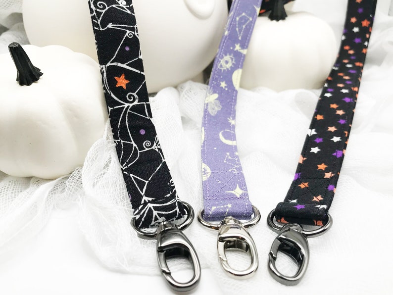 May include: Three fabric lanyards with metal swivel clasps. The lanyards feature different patterns: a black one with white spiderwebs and orange stars, a purple one with celestial designs, and a black one with orange and purple stars.