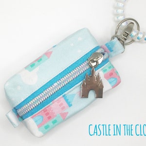 May include: A small blue zippered pouch with a white and pink castle print. The pouch has a silver zipper and a silver charm of a castle. The pouch is attached to a white and iridescent spiral keychain. The text "CASTLE IN THE CLOUDS" is printed below the pouch.