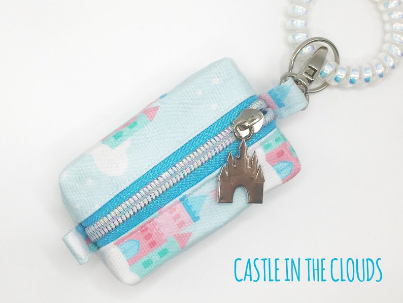 May include: A small blue zippered pouch with a white and pink castle print. The pouch has a silver zipper and a silver charm of a castle. The pouch is attached to a white and iridescent spiral keychain. The text "CASTLE IN THE CLOUDS" is printed below the pouch.