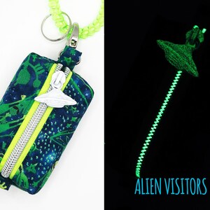 May include: A small, zippered pouch with a glow-in-the-dark zipper pull shaped like a UFO. The pouch is made of dark blue fabric with a green and white alien print. The zipper pull glows green in the dark. The text "ALIEN VISITORS" is printed in blue on a black background.