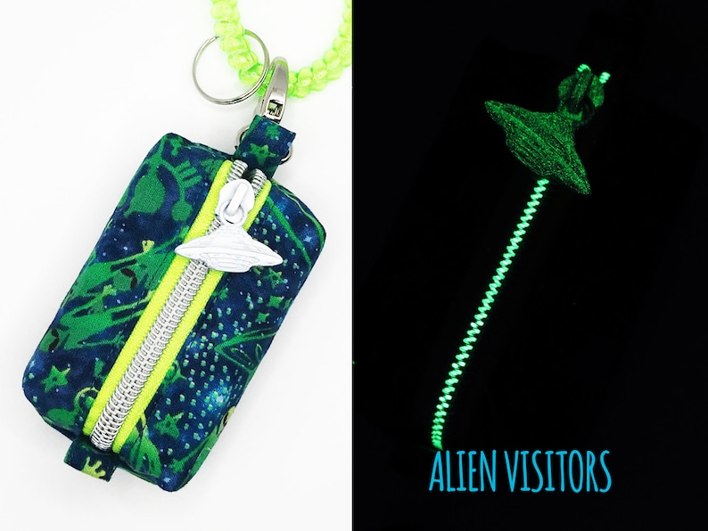 May include: A small, zippered pouch with a glow-in-the-dark zipper pull shaped like a UFO. The pouch is made of dark blue fabric with a green and white alien print. The zipper pull glows green in the dark. The text "ALIEN VISITORS" is printed in blue on a black background.