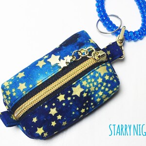 K&ouml;nnte beinhalten: Eine kleine blaue Rei&szlig;verschlusstasche mit einem goldenen Rei&szlig;verschluss und goldenen Stern-Akzenten. Die Tasche hat einen blauen und goldenen Sternenhimmel-Druck und ist an einem blauen Perlen-Schl&uuml;sselanh&auml;nger befestigt. Der Text "STARRY NIGHT" ist unter der Tasche gedruckt.