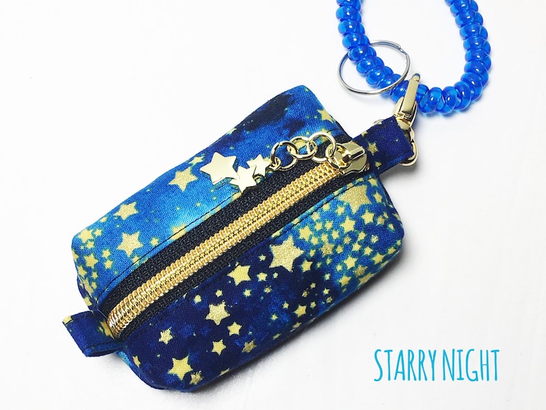 K&ouml;nnte beinhalten: Eine kleine blaue Rei&szlig;verschlusstasche mit einem goldenen Rei&szlig;verschluss und goldenen Stern-Akzenten. Die Tasche hat einen blauen und goldenen Sternenhimmel-Druck und ist an einem blauen Perlen-Schl&uuml;sselanh&auml;nger befestigt. Der Text "STARRY NIGHT" ist unter der Tasche gedruckt.