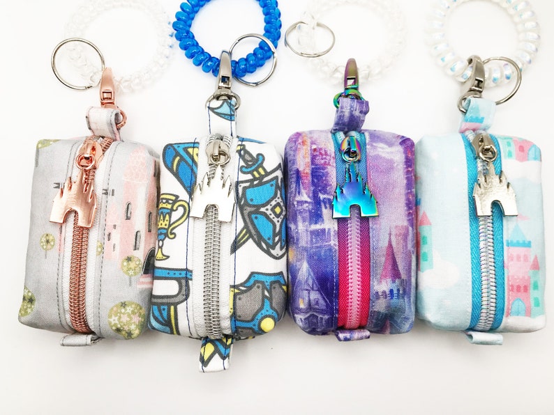 May include: Four small zippered pouches with keychains. Each pouch has a different colorful pattern and a metal charm with a castle design. The charms are in different colors, including silver, gold, and iridescent.