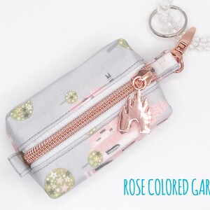 May include: A small gray zippered pouch with a pink and green floral print. The pouch has a rose gold zipper and a rose gold charm shaped like a castle. The pouch is attached to a clear beaded keychain.