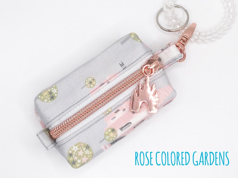 May include: A small gray zippered pouch with a pink and green floral print. The pouch has a rose gold zipper and a rose gold charm shaped like a castle. The pouch is attached to a clear beaded keychain.