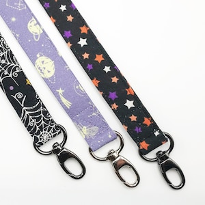 May include: Three fabric lanyards with metal swivel clasps. One is black with a white spiderweb design, another is lavender with planets and shooting stars, and the third is black with orange, purple, and white stars.