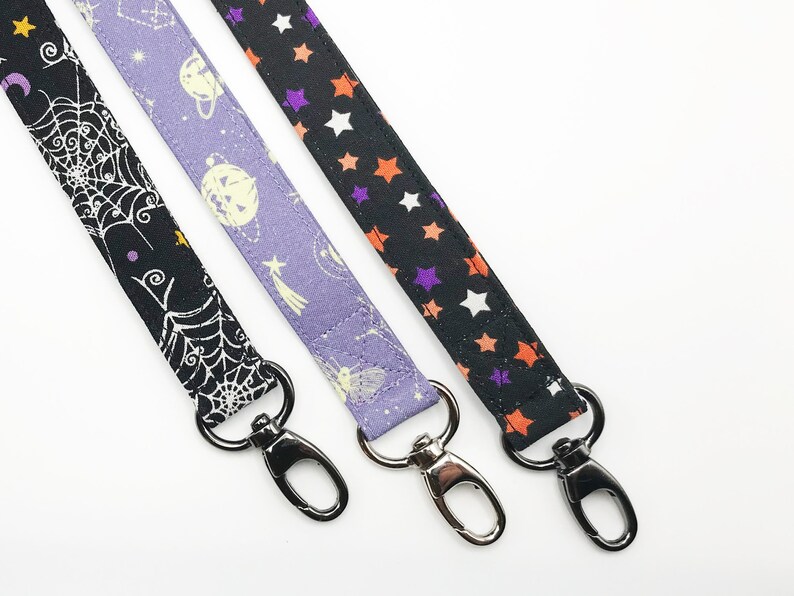 May include: Three fabric lanyards with metal swivel clasps. One is black with a white spiderweb design, another is lavender with planets and shooting stars, and the third is black with orange, purple, and white stars.