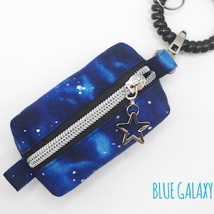 K&ouml;nnte beinhalten: Eine kleine blaue Rei&szlig;verschlusstasche mit Galaxie-Druck und einem silbernen Rei&szlig;verschluss. Die Tasche hat einen silbernen Sternanh&auml;nger am Rei&szlig;verschluss und ist an einem schwarzen Spiralschl&uuml;sselanh&auml;nger befestigt. Der Text "BLUE GALAXY" ist auf dem wei&szlig;en Hintergrund unter der Tasche gedruckt.