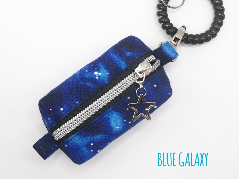 K&ouml;nnte beinhalten: Eine kleine blaue Rei&szlig;verschlusstasche mit Galaxie-Druck und einem silbernen Rei&szlig;verschluss. Die Tasche hat einen silbernen Sternanh&auml;nger am Rei&szlig;verschluss und ist an einem schwarzen Spiralschl&uuml;sselanh&auml;nger befestigt. Der Text "BLUE GALAXY" ist auf dem wei&szlig;en Hintergrund unter der Tasche gedruckt.