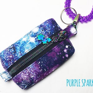 K&ouml;nnte beinhalten: Eine kleine, mit einem Galaxie-Muster in Lila und Blau bedruckte Rei&szlig;verschlusstasche mit einem silbernen Rei&szlig;verschluss und einem lila Perlen-Schl&uuml;sselanh&auml;nger. Der Text "PURPLE SPARKLES" ist auf dem wei&szlig;en Hintergrund unter der Tasche gedruckt.