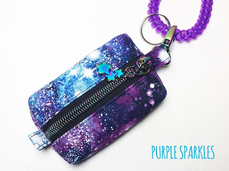 K&ouml;nnte beinhalten: Eine kleine, mit einem Galaxie-Muster in Lila und Blau bedruckte Rei&szlig;verschlusstasche mit einem silbernen Rei&szlig;verschluss und einem lila Perlen-Schl&uuml;sselanh&auml;nger. Der Text "PURPLE SPARKLES" ist auf dem wei&szlig;en Hintergrund unter der Tasche gedruckt.