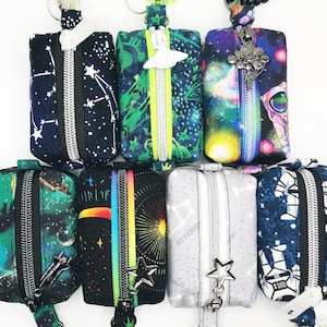 May include: Seven zippered pouches with a space theme. The pouches are made of fabric and have different designs, including stars, planets, and spaceships. The pouches are attached to key rings.