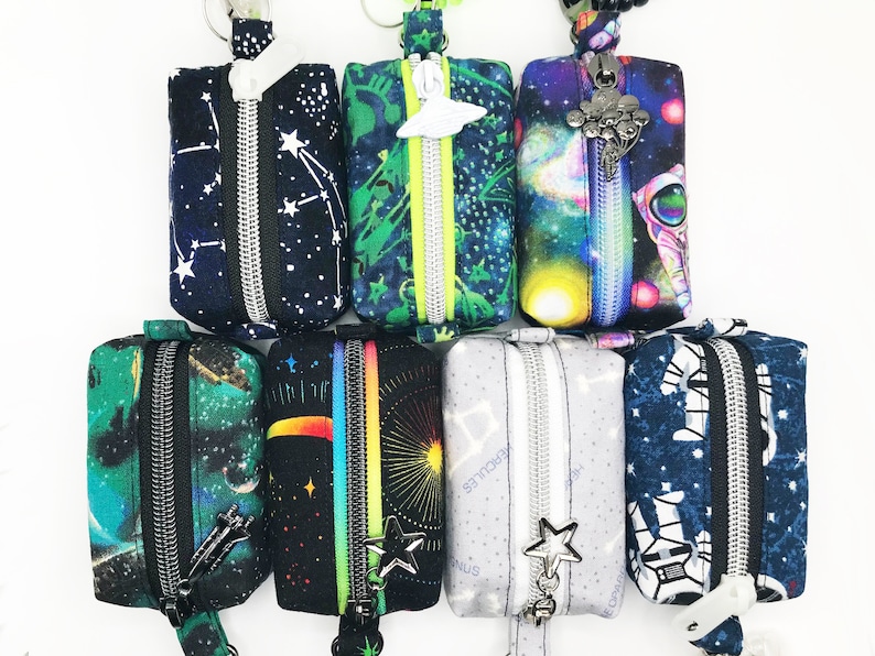 May include: Seven zippered pouches with a space theme. The pouches are made of fabric and have different designs, including stars, planets, and spaceships. The pouches are attached to key rings.