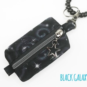 K&ouml;nnte beinhalten: Eine kleine schwarze Rei&szlig;verschlusstasche mit einem schwarzen Galaxie-Druck und einem silbernen Stern-Anh&auml;nger. Die Tasche ist an einem schwarzen Perlen-Schl&uuml;sselanh&auml;nger befestigt.