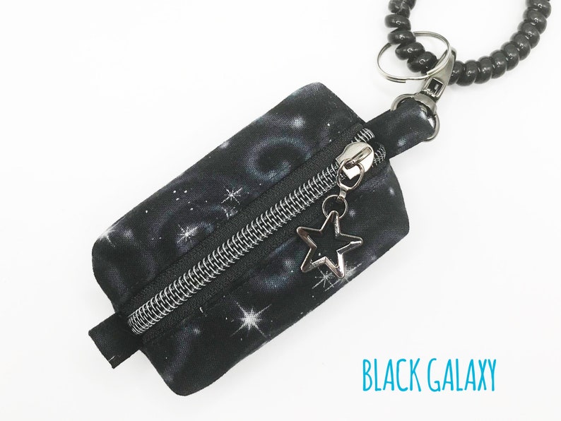 K&ouml;nnte beinhalten: Eine kleine schwarze Rei&szlig;verschlusstasche mit einem schwarzen Galaxie-Druck und einem silbernen Stern-Anh&auml;nger. Die Tasche ist an einem schwarzen Perlen-Schl&uuml;sselanh&auml;nger befestigt.