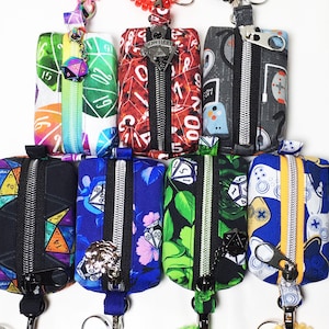 May include: Eight small zippered pouches with various colorful patterns and designs. The pouches have a zipper closure and a metal ring for attaching to a keychain. The patterns include dice, game controllers, and floral designs.