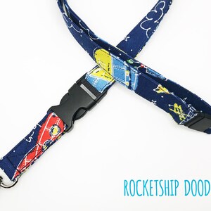 May include: A blue lanyard with a black plastic clip and a silver swivel hook. The lanyard has a pattern of rockets and stars. The text "ROCKETSHIP DOODLES" is printed on the white background below the lanyard.