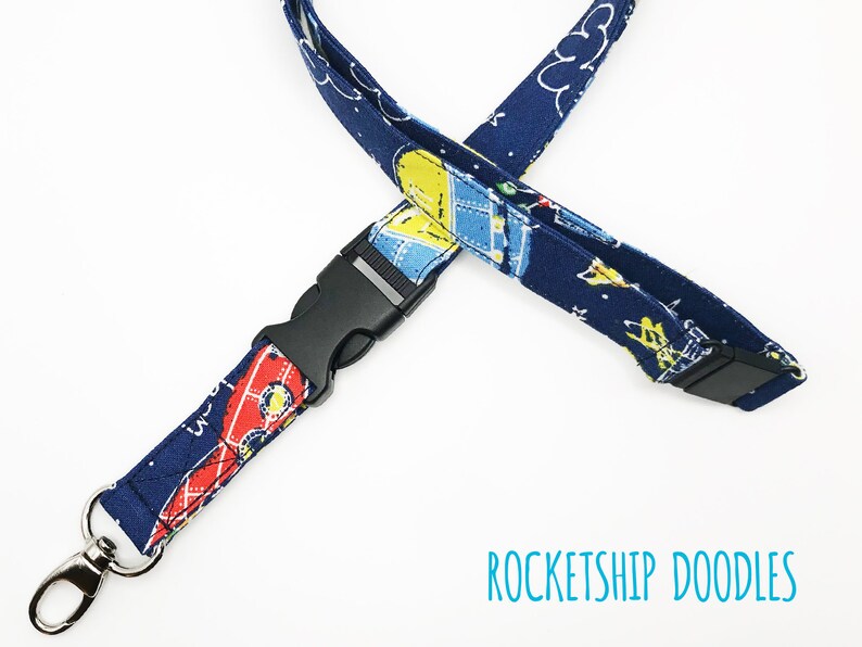 May include: A blue lanyard with a black plastic clip and a silver swivel hook. The lanyard has a pattern of rockets and stars. The text "ROCKETSHIP DOODLES" is printed on the white background below the lanyard.