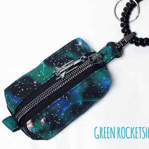 May include: A small zippered pouch with a black background and a galaxy print in shades of green and blue. The pouch has a silver zipper pull with a rocket ship charm. The pouch is attached to a black beaded key ring.