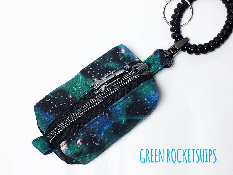 May include: A small zippered pouch with a black background and a galaxy print in shades of green and blue. The pouch has a silver zipper pull with a rocket ship charm. The pouch is attached to a black beaded key ring.