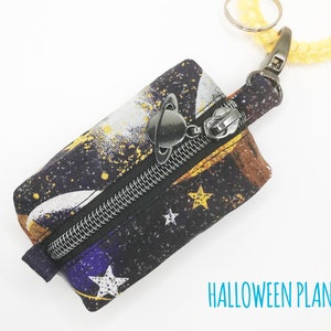 K&ouml;nnte beinhalten: Eine kleine schwarze Rei&szlig;verschlusstasche mit einem Galaxie-Druck mit Sternen, Planeten und einem wei&szlig;en Halbmond. Die Tasche hat einen silbernen Rei&szlig;verschluss und einen silbernen Ring mit einem Planeten-Anh&auml;nger, der an einem gelben Band befestigt ist.