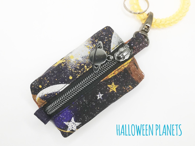 K&ouml;nnte beinhalten: Eine kleine schwarze Rei&szlig;verschlusstasche mit einem Galaxie-Druck mit Sternen, Planeten und einem wei&szlig;en Halbmond. Die Tasche hat einen silbernen Rei&szlig;verschluss und einen silbernen Ring mit einem Planeten-Anh&auml;nger, der an einem gelben Band befestigt ist.