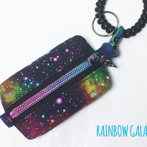 K&ouml;nnte beinhalten: Eine kleine Rei&szlig;verschlusstasche mit einem Regenbogen-Galaxie-Druck. Die Tasche ist an einem schwarzen, gewickelten Schl&uuml;sselband mit einem silbernen Ring befestigt.