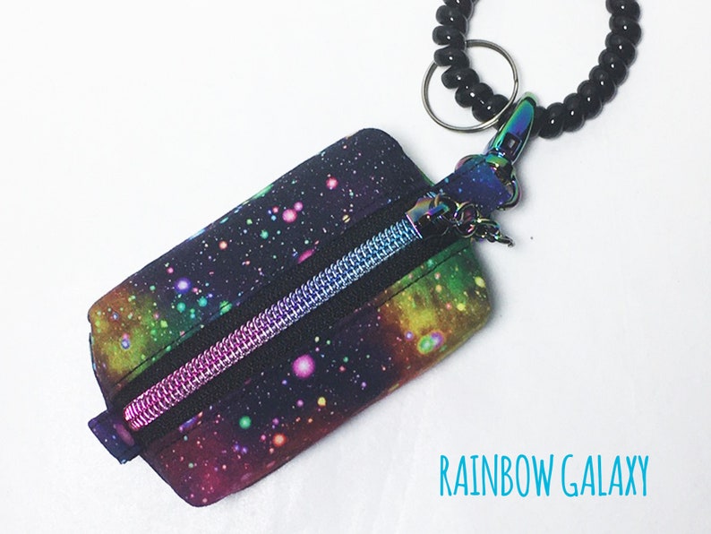 K&ouml;nnte beinhalten: Eine kleine Rei&szlig;verschlusstasche mit einem Regenbogen-Galaxie-Druck. Die Tasche ist an einem schwarzen, gewickelten Schl&uuml;sselband mit einem silbernen Ring befestigt.