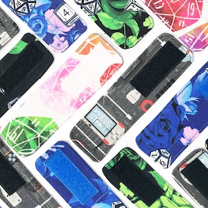 May include: Assortment of colourful fabric wristlets with various designs, including dice, flowers, and geometric patterns. Each wristlet features a black adjustable strap.