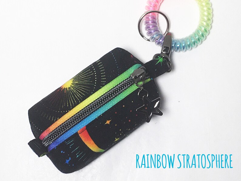 May include: A small black zippered pouch with a rainbow and star pattern. The pouch is attached to a rainbow coiled keychain with a star charm.