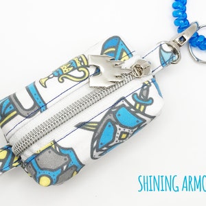 May include: A small white zippered pouch with a blue and yellow print of knights and swords. The pouch has a silver zipper and a silver charm with a crown on top. The pouch is attached to a blue coiled keychain.