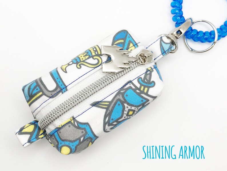 May include: A small white zippered pouch with a blue and yellow print of knights and swords. The pouch has a silver zipper and a silver charm with a crown on top. The pouch is attached to a blue coiled keychain.