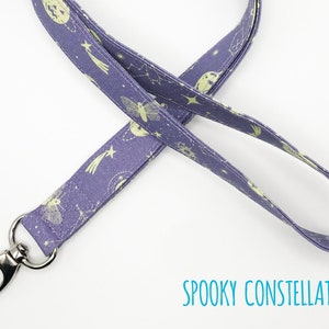 May include: A purple lanyard with a repeating pattern of celestial bodies and a silver-colored metal clasp. The lanyard features a design of moons, stars, and constellations. The text "SPOOKY CONSTELLATION" is printed in teal.