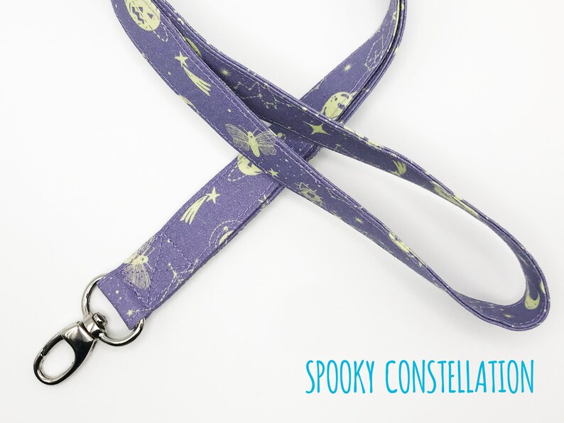 May include: A purple lanyard with a repeating pattern of celestial bodies and a silver-colored metal clasp. The lanyard features a design of moons, stars, and constellations. The text "SPOOKY CONSTELLATION" is printed in teal.
