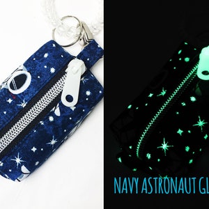 May include: A small blue zippered pouch with a white zipper pull and a white astronaut design. The pouch is decorated with white stars and glows in the dark. The text "NAVY ASTRONAUT GLOW" is printed below the pouch.
