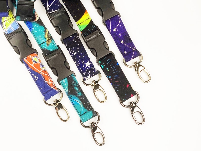 May include: Five colorful lanyards with black plastic clips and silver swivel clasps. The lanyards feature a space-themed print with stars, planets, and constellations.