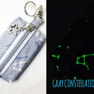 May include: A small gray zippered pouch with a silver star charm and a white zipper. The pouch is printed with white constellations and the names of the constellations in blue. The pouch glows green in the dark.