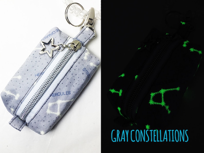 May include: A small gray zippered pouch with a silver star charm and a white zipper. The pouch is printed with white constellations and the names of the constellations in blue. The pouch glows green in the dark.
