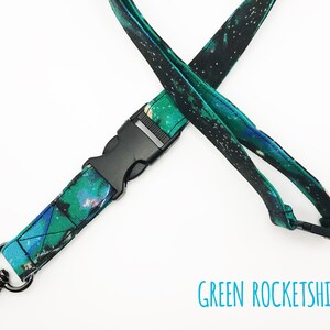May include: A green and black lanyard with a galaxy print and a black plastic clip. The lanyard has a silver metal hook on one end. The text "GREEN ROCKETSHIPS" is printed below the lanyard.