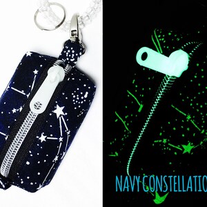 May include: A small zippered pouch with a navy blue background and white constellations. The pouch has a silver zipper and a white zipper pull. The pouch is attached to a silver key ring. The pouch glows green in the dark. "NAVY CONSTELLATIONS" is printed on the pouch.