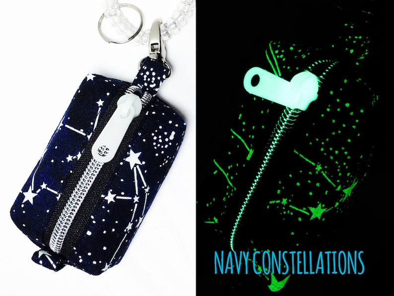 May include: A small zippered pouch with a navy blue background and white constellations. The pouch has a silver zipper and a white zipper pull. The pouch is attached to a silver key ring. The pouch glows green in the dark. "NAVY CONSTELLATIONS" is printed on the pouch.