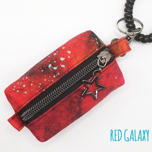 K&ouml;nnte beinhalten: Eine kleine rote Rei&szlig;verschlusstasche mit Galaxie-Druck und einem schwarzen Rei&szlig;verschluss. Die Tasche ist an einem schwarzen Spiralkettchen mit einem silbernen Sternanh&auml;nger befestigt. Der Text "RED GALAXY" ist unter der Tasche aufgedruckt.