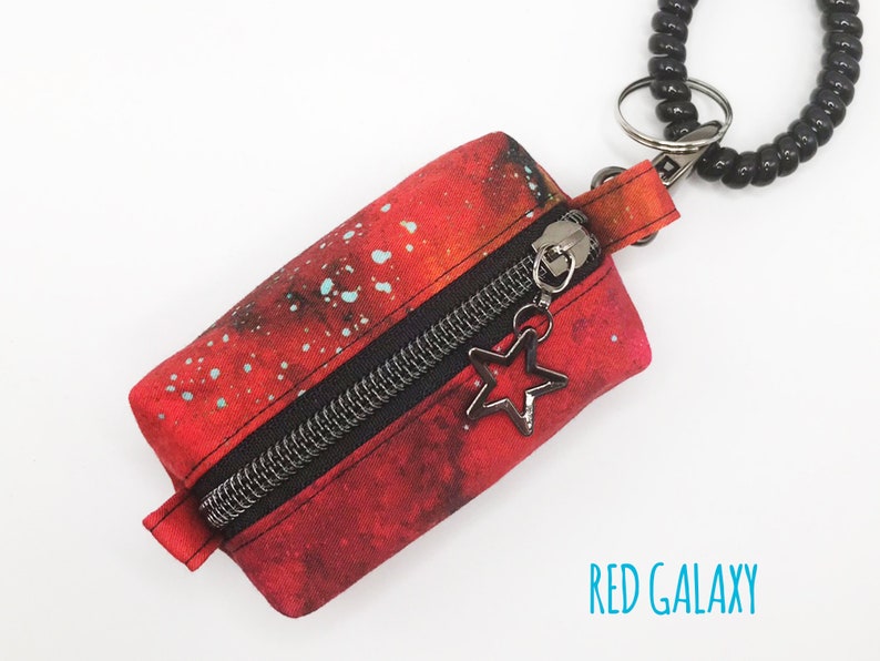 K&ouml;nnte beinhalten: Eine kleine rote Rei&szlig;verschlusstasche mit Galaxie-Druck und einem schwarzen Rei&szlig;verschluss. Die Tasche ist an einem schwarzen Spiralkettchen mit einem silbernen Sternanh&auml;nger befestigt. Der Text "RED GALAXY" ist unter der Tasche aufgedruckt.