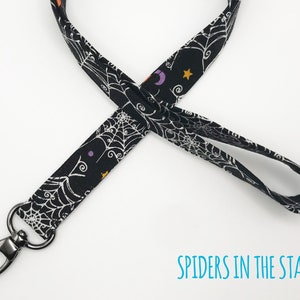 May include: Black lanyard with a Halloween theme. The fabric features a white spiderweb design, along with purple crescent moons, orange stars, and other small shapes. The lanyard has a silver-colored swivel clasp and the text "SPIDERS IN THE STARS".