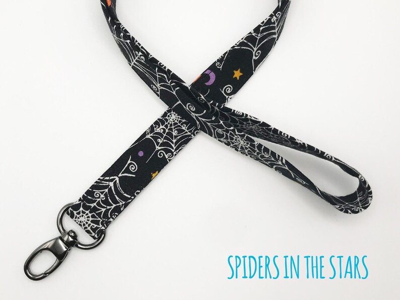 May include: Black lanyard with a Halloween theme. The fabric features a white spiderweb design, along with purple crescent moons, orange stars, and other small shapes. The lanyard has a silver-colored swivel clasp and the text "SPIDERS IN THE STARS".